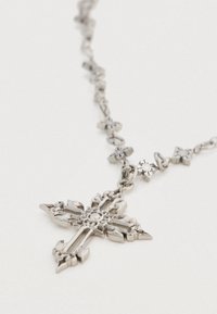 Ασημένιο μεταλλικό κολιέ με μεγάλο, περίτεχνο μενταγιόν σε σχήμα σταυρού. Ο σχεδιασμός έχει περίπλοκες λεπτομέρειες και μικρούς συνδέσμους σε σχήμα σταυρού στην αλυσίδα.