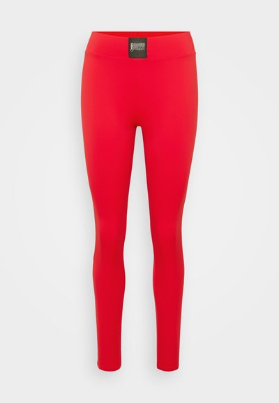 Leggings vermelhos feitos de um tecido liso e elástico, com uma cintura alta e uma marcação subtil na parte da frente. Não são visíveis costuras.