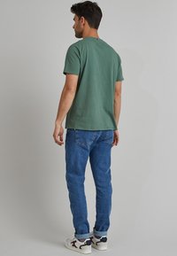 T-shirt di cotone verde con scollo rotondo, maniche corte e un design semplice, abbinata a jeans di denim blu e sneakers bianche.