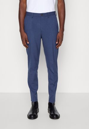 Mann trägt schmal geschnittene blaue Anzughose, schwarze Lederknöchelstiefel und ein weißes Hemd, steht vor einem hellgrauen Hintergrund.