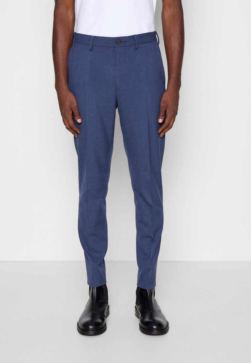Homme portant un pantalon habillé bleu slim, des bottines en cuir noires et une chemise blanche, debout devant un fond gris clair.