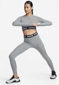 Conjunto atlético gris que incluye una camiseta de manga larga y leggings de cintura alta. La pretina tiene un logo negro de "NIKE PRO". Tejido texturizado con acentos suaves.
