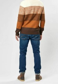 Personne portant un pull rayé marron, beige et orange, un jean bleu et des bottes marron, debout de dos sur un fond uni.