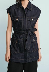Dżinsowa koszula bez rękawów z dwoma kieszeniami na piersi, srebrnymi okuciami i ozdobą w postaci wiązania w talii. Ciemny odcień z kontrastowym przeszyciem.