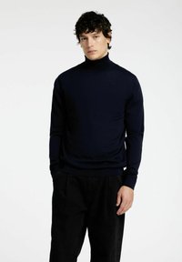 Mornarsko pulover s turtleneck iz lahkega materiala in prilagojeno obliko. Ima dolge rokave in rebraste manšete, v kombinaciji s črnimi hlačami.