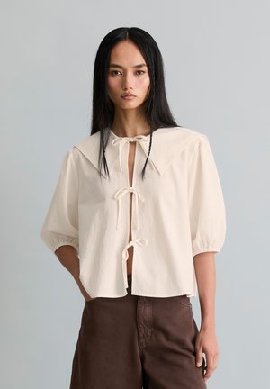 Femme aux longs cheveux noirs portant un chemisier crème avec un grand col et des fermetures à nouer, associé à un pantalon marron taille haute.
