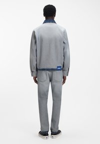 Chaqueta y pantalones de mezclilla en gris claro, con un cuello azul oscuro, detalle de logo azul y diseño de pierna recta. Textura suave y ajuste casual.