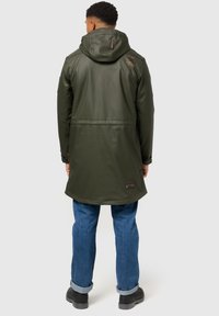 Olive green waterproof jacket with a hood, featuring a zip closure, adjustable cuffs, and a back vent. Worn with blue denim jeans and black boots.