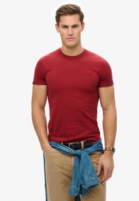 Camiseta de algodón roja con mangas cortas, cuello redondo y un pequeño logo en el pecho. Combinada con pantalones beige y una camisa de mezclilla azul atada a la cintura.