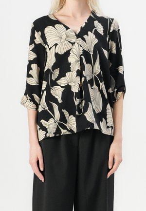 Blouse noire à grand motif floral beige et manches trois-quarts portée avec un pantalon noir à jambes larges.