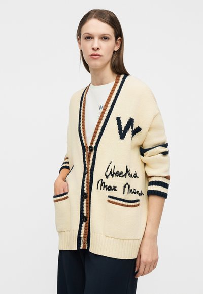 Γυναίκα που φοράει εκρού πλεκτή ζακέτα με μαύρες και καφέ ρίγες, κλείσιμο με κουμπιά, μπροστινές τσέπες και κέντημα με κείμενο πάνω από λευκό πουκάμισο και σκούρο παντελόνι.