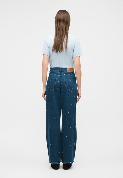 Γυναίκα που στέκεται γυρισμένη προς τα πίσω, φοράει ανοιχτό γαλάζιο κοντομάνικο τοπ και ψηλόμεσο, φαρδύ, σκούρο μπλε τζιν με λεπτά μικρά λευκά σχέδια.