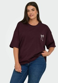 Bordeaux oversized t-skjorte laget av bomull, med korte ermer og en hvit brodert sløyfe detalj på brystet. Kombinert med blå jeans.