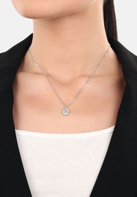 Collar de péndulo de brújula plateado llevado por una mujer con un blazer negro y una blusa blanca, recortado por debajo de la barbilla sobre un fondo blanco.