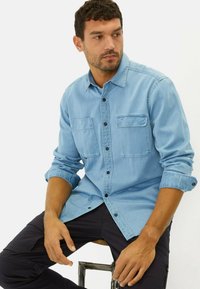 Lichtblauw denim shirt met lange mouwen, knoopsluiting, twee borstzakken en zwarte knopen, gestyled over donkere broeken.