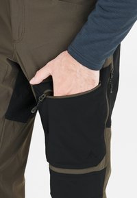 Hånd, der stikker ind i sort lynlåslomme på låret på tofarvede brune og sorte udendørsbukser.