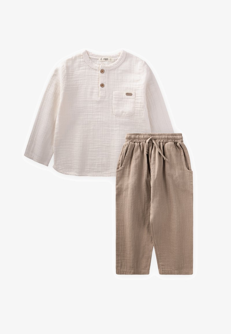 Duga bijela pamučna košulja s džepom na prsima i dva gumba, uparen s bež hlačama s elastičnim strukom i bočnim džepovima.