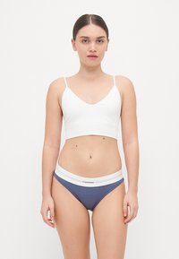 Hvit spandex bralette med tynne stropper, kombinert med marineblå ribbestrikkede bikiniunderdeler med hvit midjebånd og 'Calvin Klein'-logo.