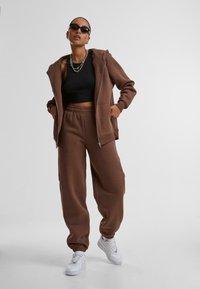 Bruine rits-hoodie met kangaroo zakken en bijpassende sweatpants. Zwarte cropped top en witte sneakers met veters. Zonnebril en kettinghalsband.