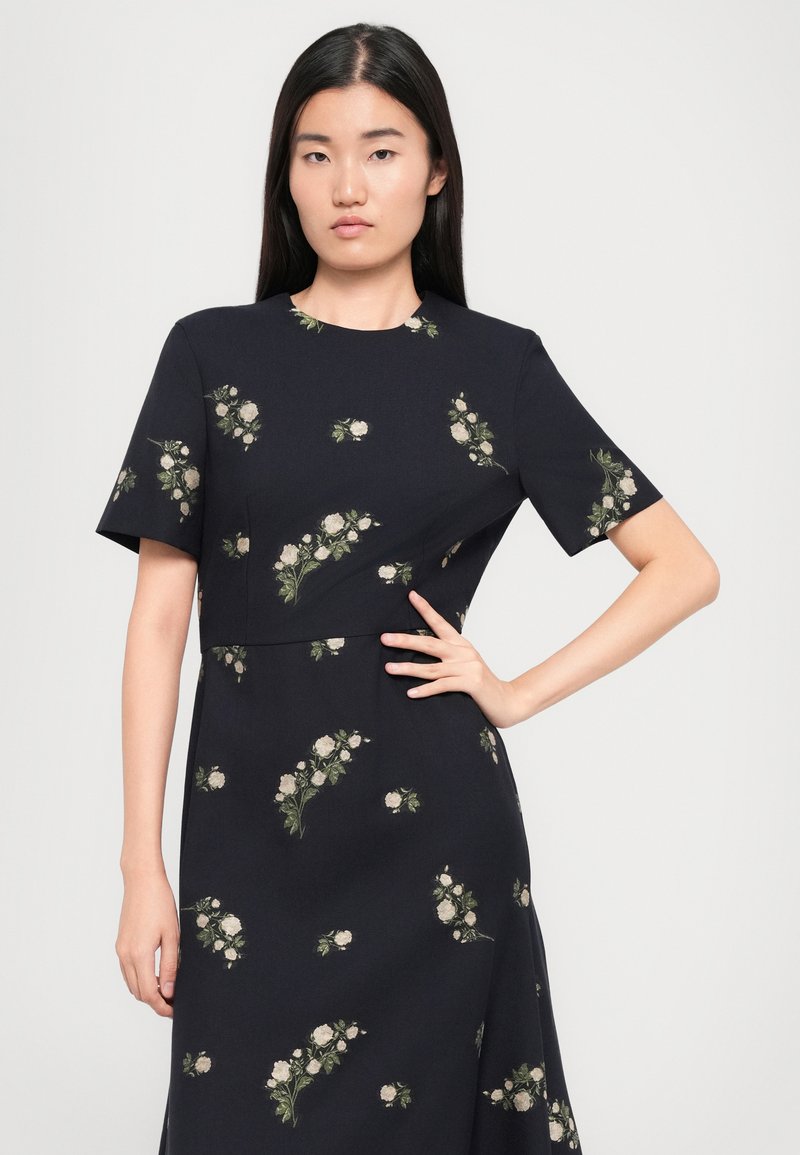 Robe de couleur marine avec des manches courtes et un motif floral en crème et vert. Le corsage ajusté se prolonge en une jupe évasée. Texture de tissu lisse.