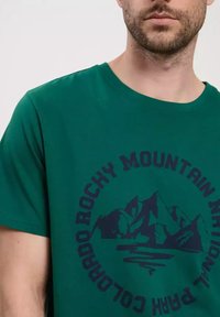 Zielona bawełniana koszulka z okrągłym dekoltem i krótkimi rękawami. Na przedniej stronie grafika przedstawiająca góry oraz tekst w granatowym kolorze.