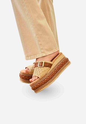Pies que llevan sandalias de plataforma beige con textura tejida y correas de hebilla de cuero, combinadas con pantalones color beige claro sobre un fondo liso.