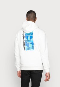 Vit hoodie med en blå grafik på baksidan som har vågmönster och texten "adidas originals" i svart. Designad med en kenguruficka och justerbar huva.
