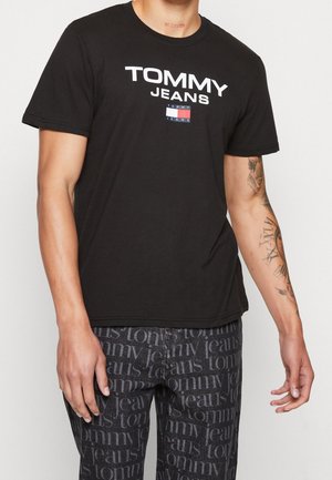 Mężczyzna ubrany w czarną koszulkę Tommy Jeans i ciemne spodnie z powtarzającym się napisem "tommy jeans", ukazujący tatuaż na prawym przedramieniu i dłoni.