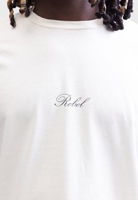 Weißes Baumwoll-T-Shirt mit dem Schriftzug "Rebel" in schwarzer Schreibschrift auf der Vorderseite, klassischem Rundhals und glatter Textur.