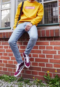 Gul sweatshirt med en röd Levi's-logotyp, ljusblå jeans och vinröda sneakers med vita snören, lutande mot en tegelvägg.