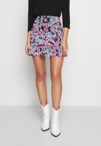 Femme portant un haut noir, une mini-jupe florale avec des fleurs roses et bleues, et des bottes de cowboy blanches mi-mollet à bouts pointus, se tenant devant un fond uni.