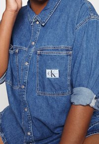 Henkilö, joka käyttää käännetyillä hihoilla olevaa sinistä Calvin Klein farkkupaitaa, jossa on näkyvä merkki rinnustaskussa.