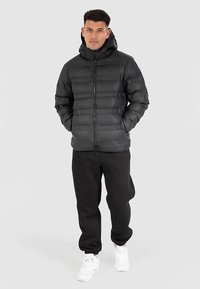 Sort puffjakke med lynlåslukning foran, hætte og quiltet design, kombineret med sorte joggingbukser og hvide sneakers.