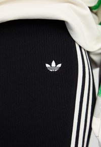 Čierne rebrované nohavice s tromi bielymi vertikálnymi pruhmi po boku; majú malé biele logo Adidas na stehne; mäkká textúra látky.