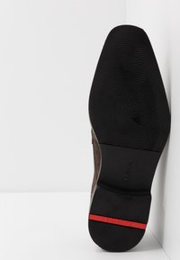 Semelle en suède marron avec un fond en caoutchouc noir texturé, comportant une bande d'accent rouge et une semelle intermédiaire lisse.