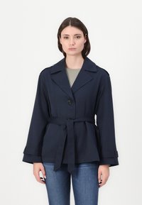 Marineblauwe riemjas gemaakt van gladde stof, met een opstaande kraag, twee voorzakken en knoopsluiting. Gestyled met brede mouwen.