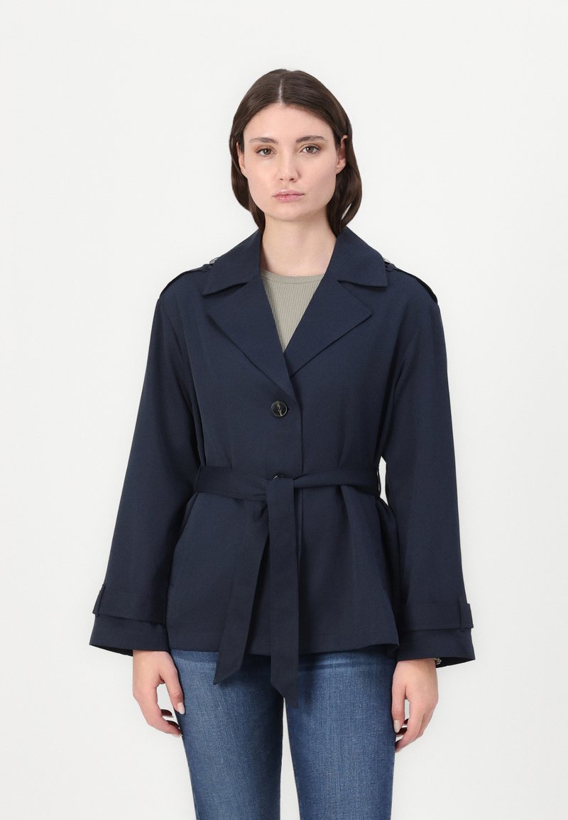 Marineblauwe riemjas gemaakt van gladde stof, met een opstaande kraag, twee voorzakken en knoopsluiting. Gestyled met brede mouwen.