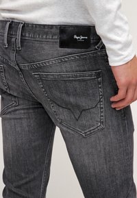Osoba v bledých černých džínech Pepe Jeans s výraznou vyšitou zadní kapsou a koženou nášivkou značky, ruka opřená u kyčle.