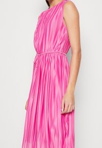 Różowa plisowana sukienka z okrągłym dekoltem i bezrękawowym fasonem. Posiada wcięcie w talii z przewiązanym paskiem oraz lekki, zwiewny materiał.