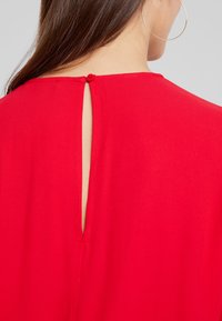 Top rojo con abertura en forma de llave en la parte trasera, asegurada con un pequeño botón. Tela suave con un diseño estructurado.