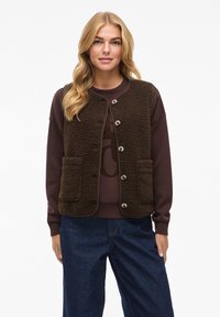 Braune, strukturierte Fleece-Weste mit Rundhalsausschnitt und vorderen Knöpfen. Verfügt über zwei vordere Taschen. Getragen über einem dunklen Pullover und blauen Hosen.