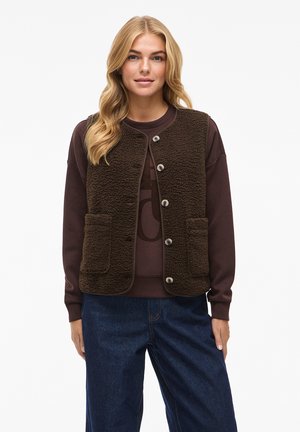 Bruine, textuur fleece vest met ronde hals en knopen aan de voorkant. Beschikt over twee voorzakken. Draag het over een donkere trui en blauwe broek.