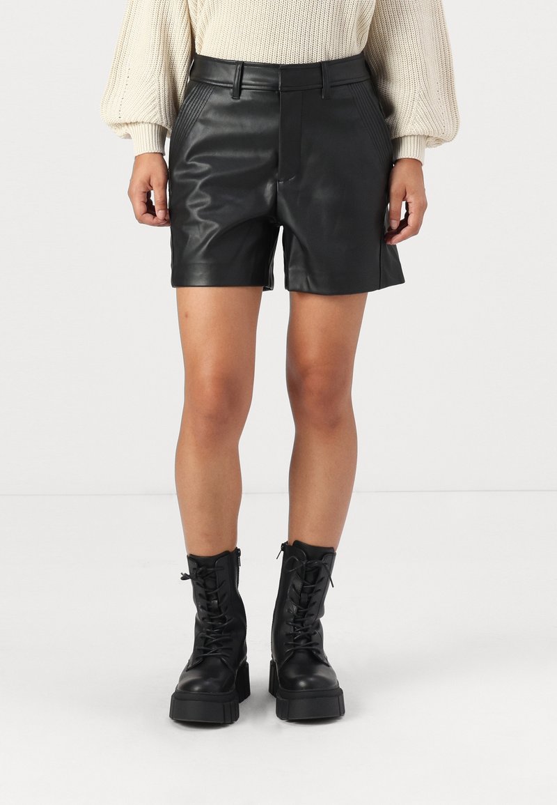 Personne portant un short en cuir noir, un pull en tricot crème et des bottes plateformes à lacets noires, debout devant un fond blanc.