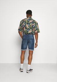 Camisa de manga corta de color azul marino con estampado floral en verde y rosa, combinada con pantalones cortos de mezclilla enrollados. El modelo lleva zapatillas negras y calcetines blancos.