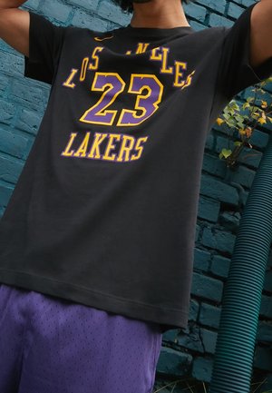 Osoba má na sobě černé tričko Los Angeles Lakers s číslem 23 ve žluté a fialové barvě a fialové sportovní šortky, stojící opřená o modrou cihlovou zeď.