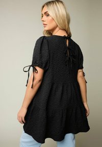 Blusa nera in tessuto testurizzato con maniche a sbuffo, caratterizzata da un design a strati e lacci posteriori. Il tessuto ha un aspetto stropicciato, offrendo una vestibilità rilassata.