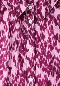Korte mouw shirt in een roze en donkerpaars ikatpatroon. Voorzien van een puntige kraag en vijf witte knopen aan de voorkant.