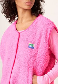 Flis brezrokavnik v svetlo rožnati barvi z teksturirano površino in kontrastnim robom. Ima logotipno značko, prek katerega je oblečena rožnata majica z dolgimi rokavi.