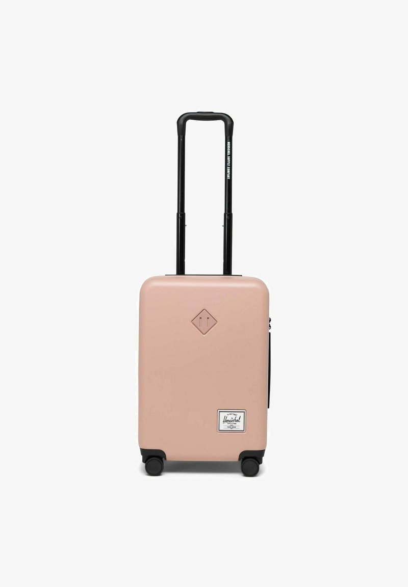 Valise rigide compacte en rose clair mat avec accents noirs, poignée télescopique et quatre roues pour une mobilité facile. Comprend un patch de logo.