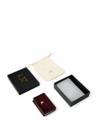 Portfel z bordowej skóry z wzorem krokodyla i złotym akcentem, umieszczony obok czarnego pudełka upominkowego i beżowego woreczka ze sznurkiem.